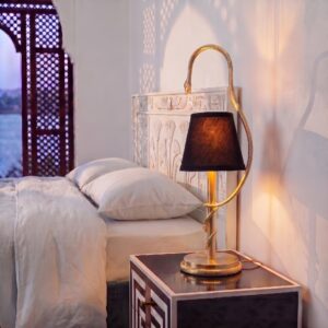 Cleo serpent table lamp in unlacquered brass on a nightstand beside a bed with an Egyptian-carved headboard